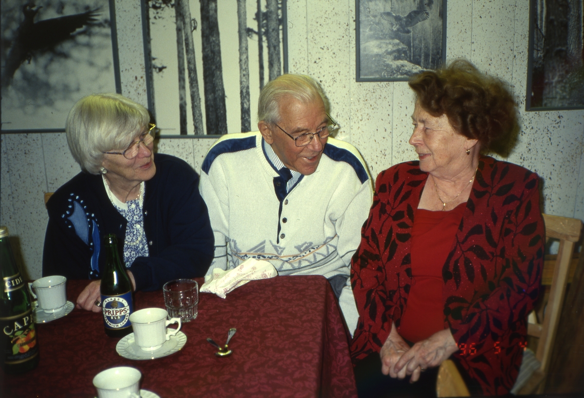 Adéle(till höger i bild) sitter vid ett bord och samtalar med en man och en kvinna. På bordet syns både en ölflaska och en ciderflaska samt tre kaffekoppar. På väggen bakom hänger inramade fotografier av fåglar som Hilding tagit.