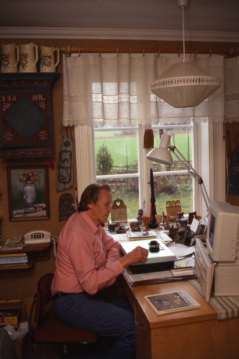 Hilding Mickelsson sitter vid sitt skrivbord i arbetsrummet och tittar på negativ genom ett förstoringsglas. På skrivbordet står en dator.