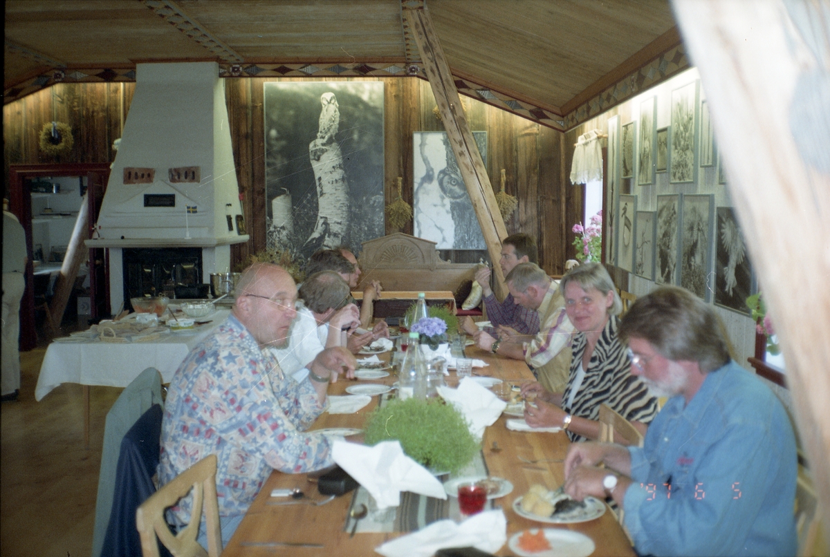 En grupp människor sitter och äter vid ett bord, på övervåningen till en av byggnaderna på Sunnanåker 5 juni 1994. Hildings bilder pryder väggarna.