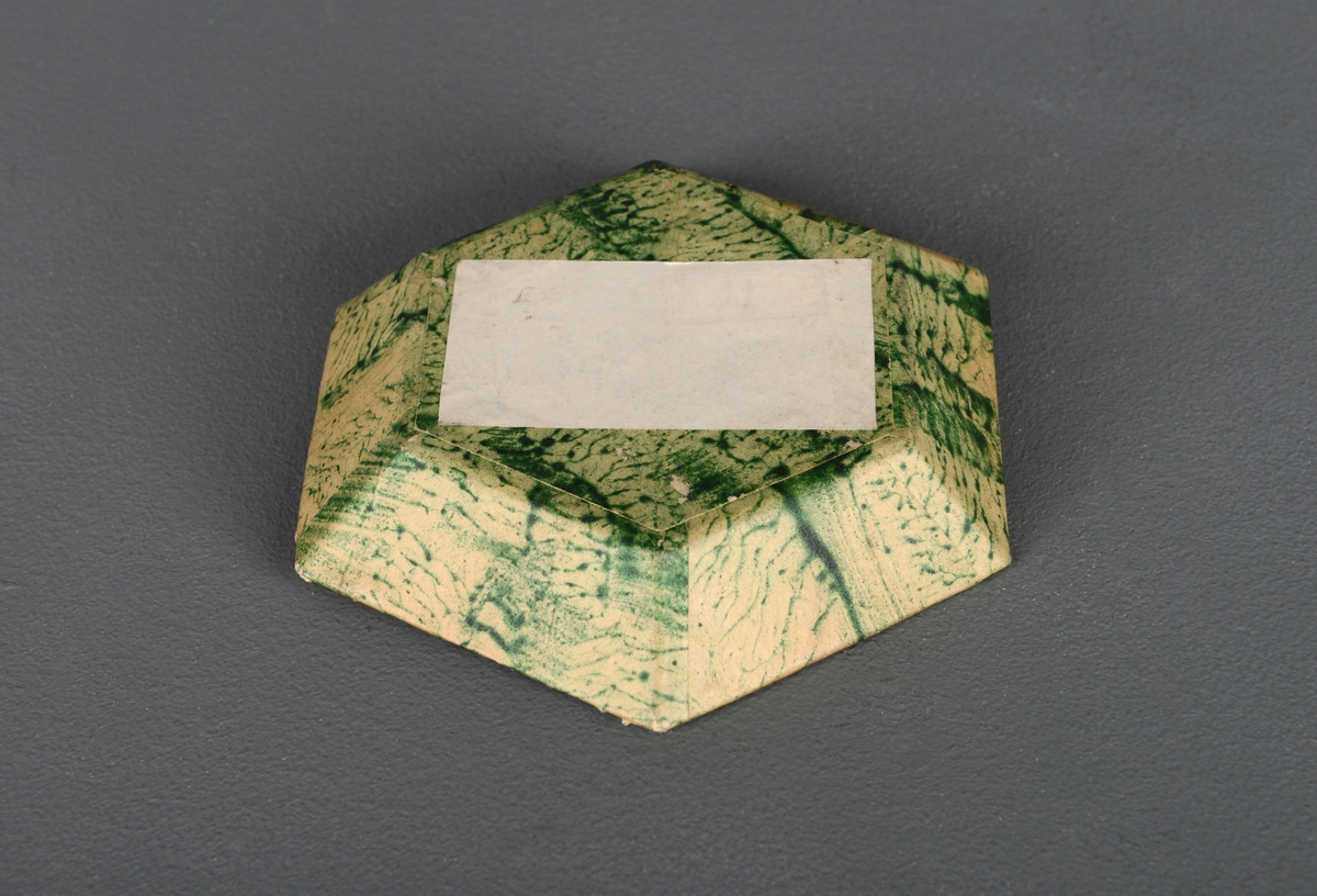Et lite sekskantet fat, laget i papp og trekt med papir. Papiret har marmorert mønster, og på innsiden er det brunt og blågrønt mønster, mens på utsiden er den beige og grønn. På undersiden er det festet en rektangulær papirlapp. Laget i pappsløyd.