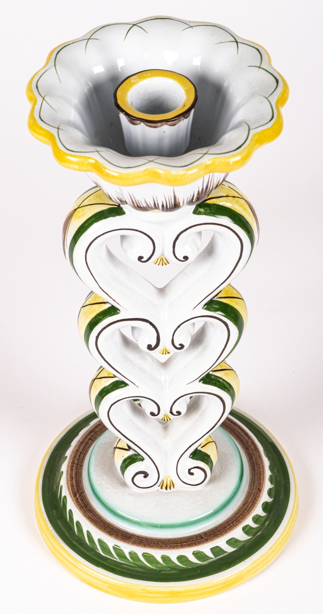 Ljusstake modell H från Gefle Porslinsfabrik, formgiven av konstnär Arthur Percy 1926 för fabriken. Rund fotplatta som bär upp tre på varandra stående stiliserade hjärtan, överst en ljushållare i form av en öppen blomma. Ljusstake H har tillverkats med olika glasyrer. Just denna är handmålad i dekor 64 av chefsdekoratör Eugen Trost, som signerat med T. på undersidan.