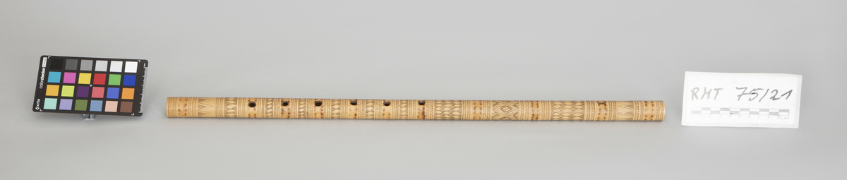 Sylindrisk fløyte av bambus i et stykke. Dekorert med innrissete stiliserte border og brennemerker, seks fingerhull brent inn i røret.