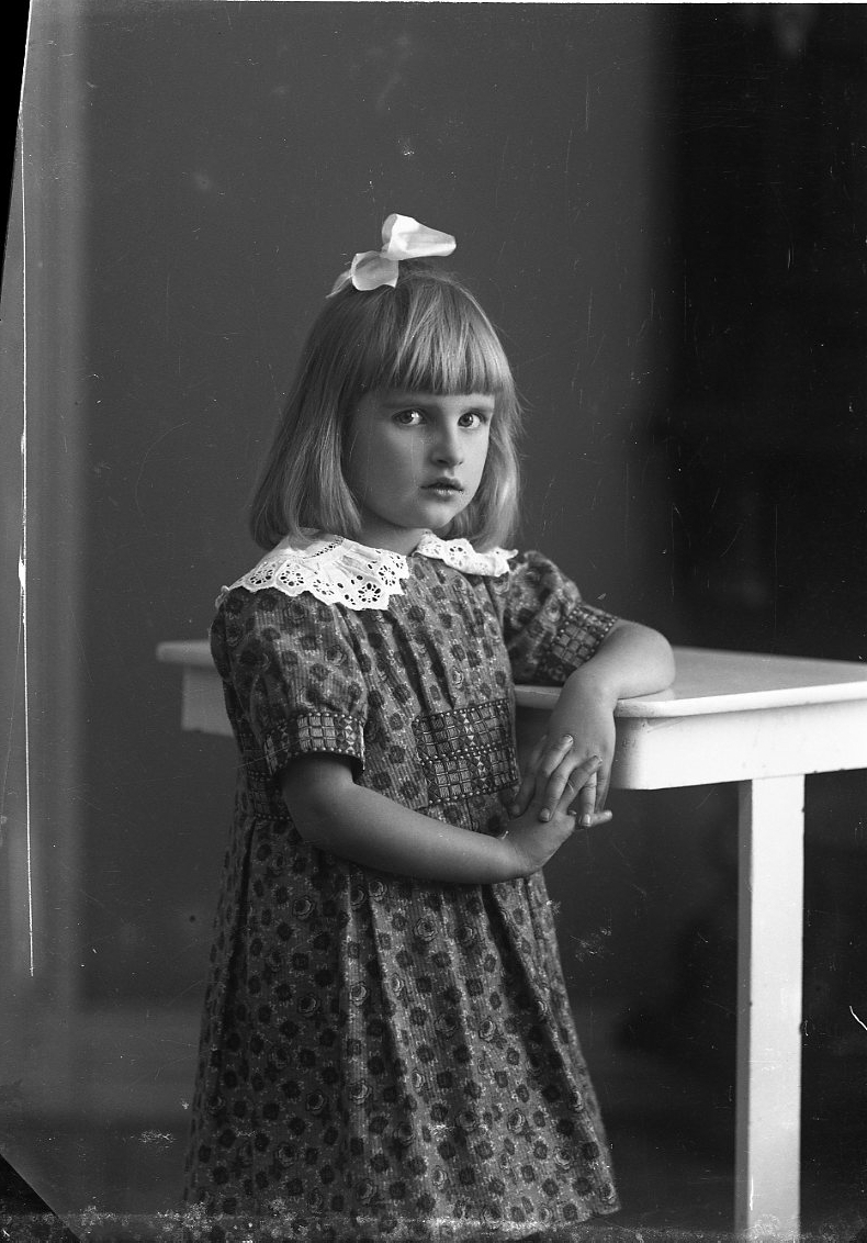 Porträtt av en flicka i prickig klänning med spetskrage och rosett i håret. Hon står lutad mot ett bord. Hennes efternamn är Witt.