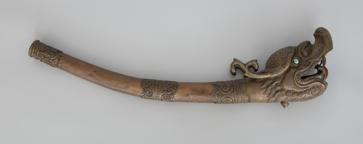 Blåseinstrument av kobber, med munnstykke, tverrbånd og klokkestykke av ornamentert messing. Klokkestykket har form av et dragehode med oppsperret gap. Øyer av turkis edelsten.