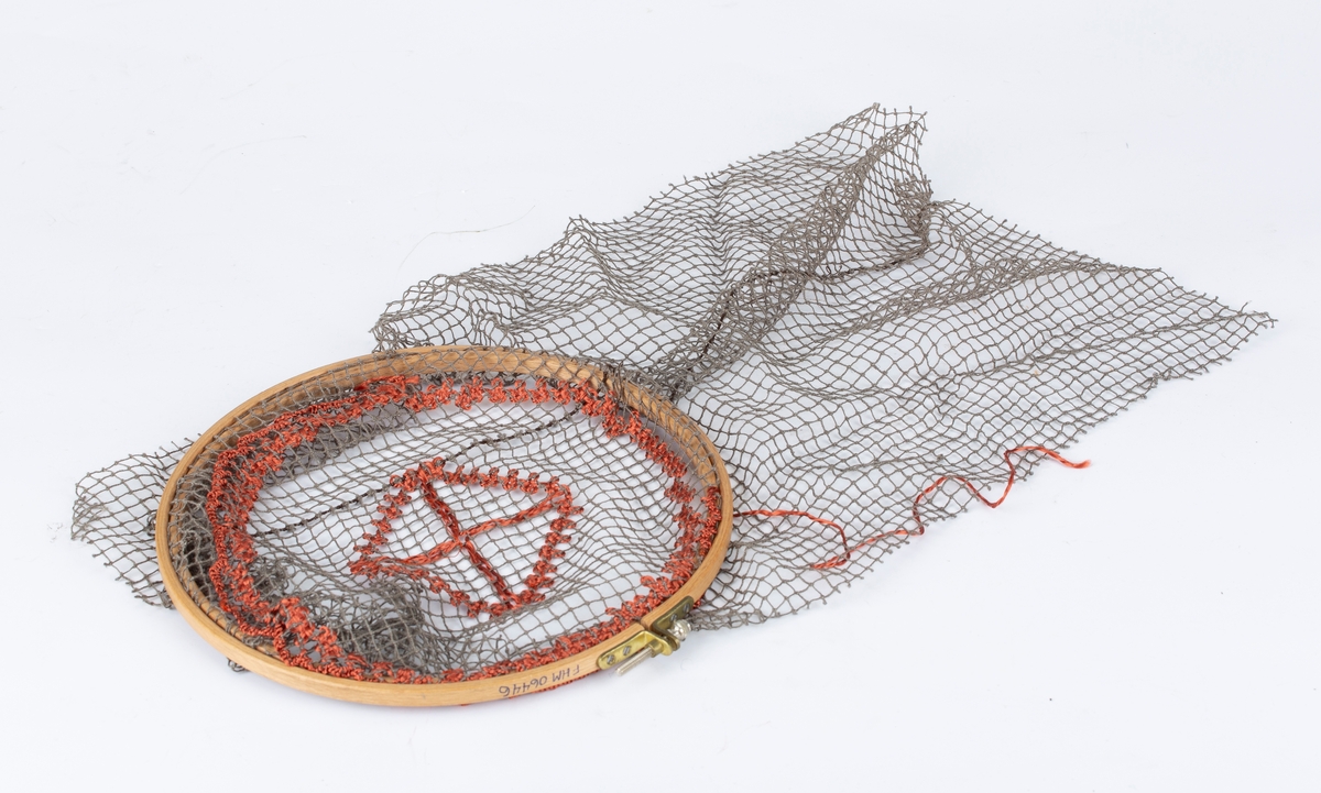 Sirkelrund håndarbeidsramme, påspent nettingbunn for fileringsarbeid. Påbegynt mønster med rustrød tråd (tvunnet silketråd?).