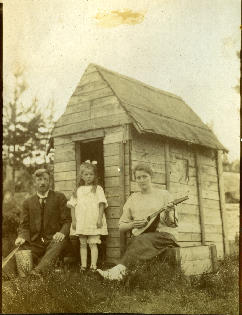 En mann, en jente og en kvinne med mandolin foran et lekehus.