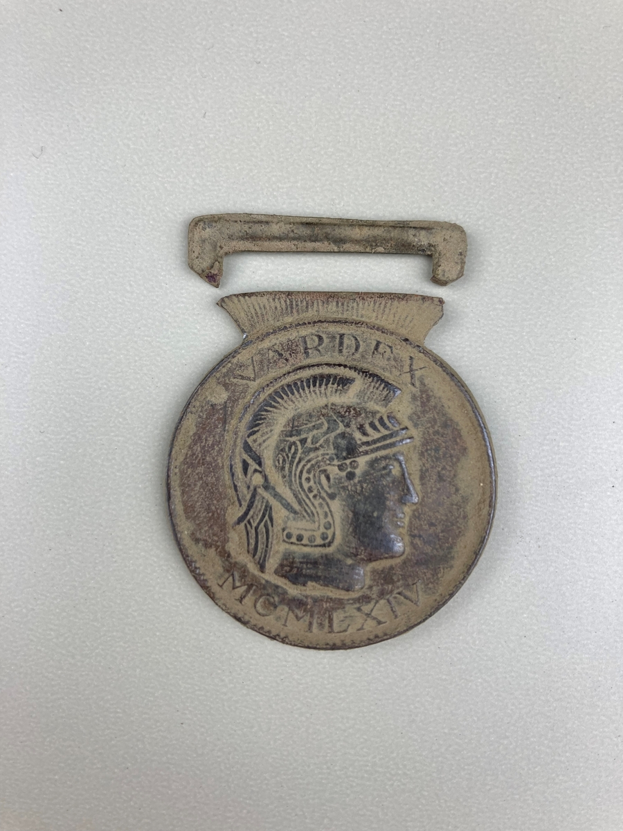 Rund gjenstand i metall, ligner en medalje. Markert "V A R D E/F X", " M O M L X I V" (1969?).
