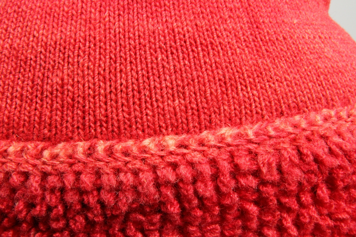 Rød strikket topplue. Lua er tett strikket med små pinner og forholdsvis tynt, hardt spunnet garn. Med flosset 8,5 cm bred kant nederst, ant. strikket i løkkestrikk. Liten dusk på toppen. Sammensydd i spissen. Foret med naturhvit strikket for. 
