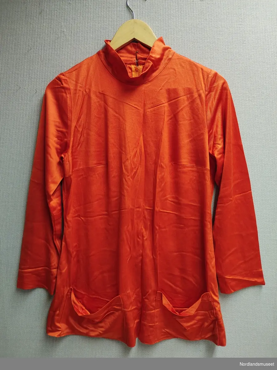 Høyhalset bluse med to lommer på framsiden og lange ermer. 
Blusen har to hemper og glidelås i ryggen. 
Sydd i et syntetisk stoff.