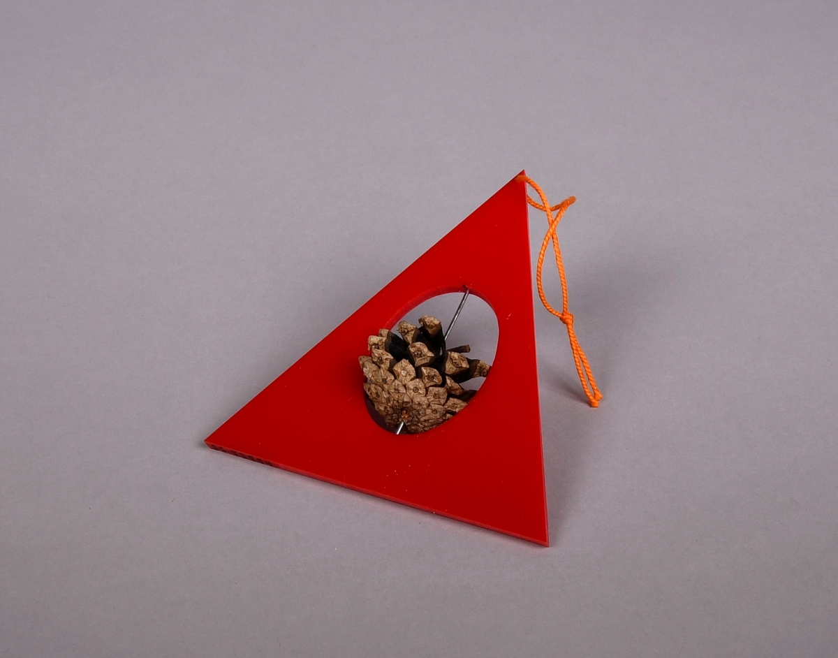 Juletrefigur av farget akryl og furukongle. Juletre formet som rødfarget trekant med ovalt hull i midten, i hullet er det en stav av metall som går gjennom en furukongle.