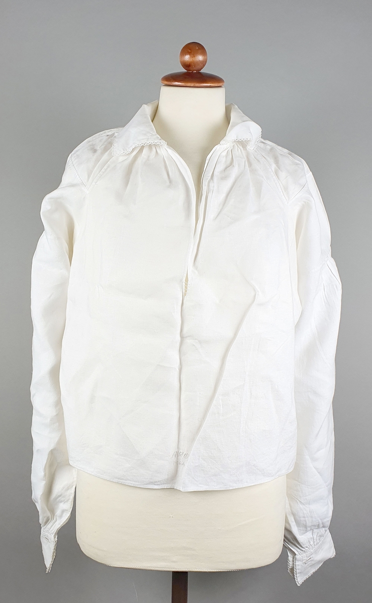 Hvit bunadskjorte av lin, med krage og mansjetter med nupereller. Rysjer langs halslinningen. Skjorten er åpen i brystet, men ikke helt ned.