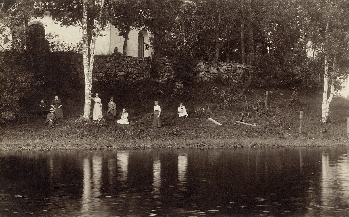Ett sällskap har slagit sig ner vid strandkanten av en sjö, med en kyrka på höjden bakom. Ca 1912.
Stenbrohult (?), Ljuders kyrka (?, före sjösänkningen).