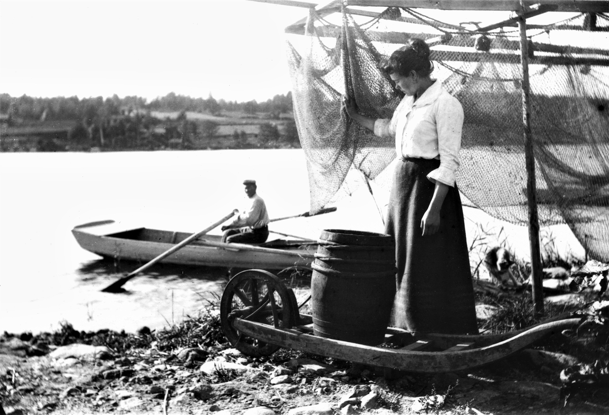 Man i roddbåt vid stranden där en kvinna står bakom ett upphängt fiskenät. En tunna står på en låg kärra framför henne.