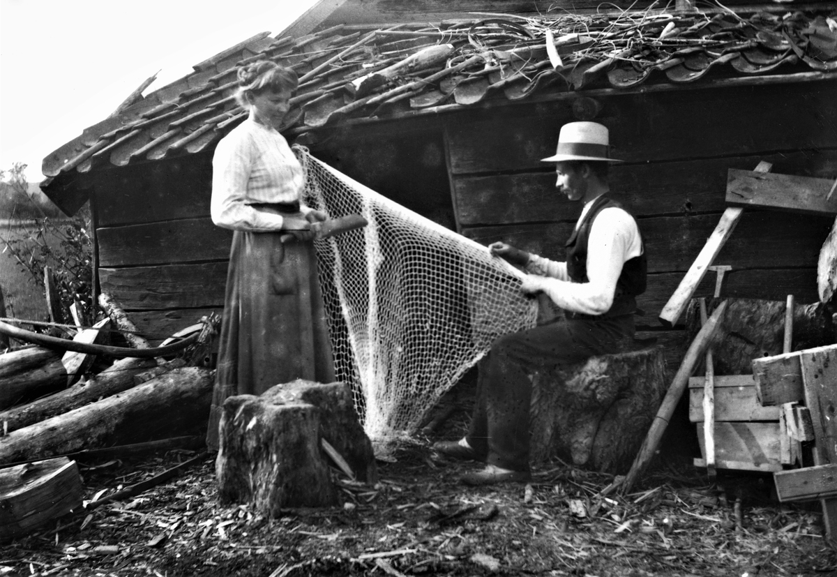 Kvinna och man lagar nät på vedbacken. Boden har enkupigt taktegel.