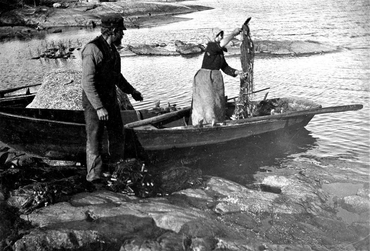 Ett äldre par i sin fiskebåt lastar ur garnen i en havsvik.
(Se även bildnr VMA6718_6)