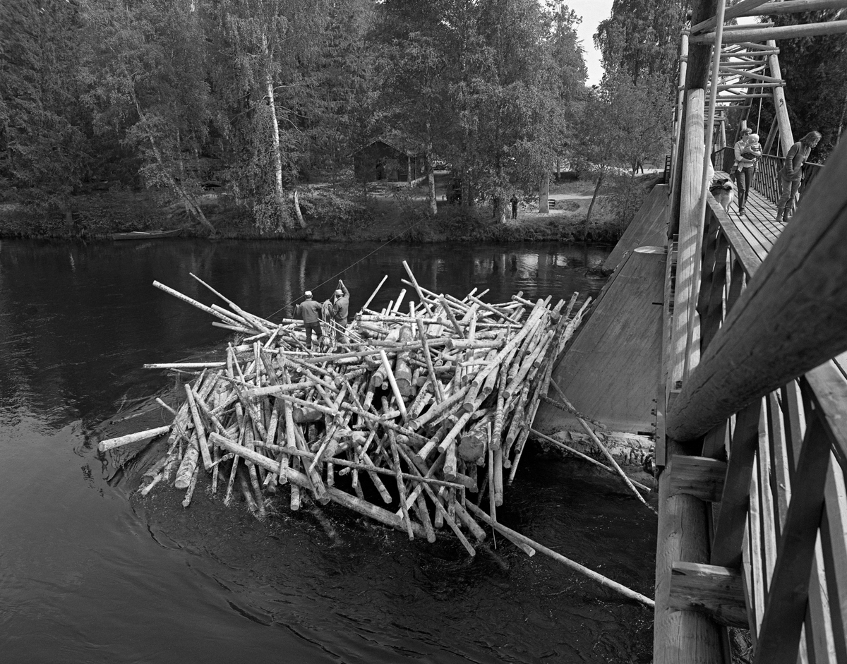 Tømmerhauger på motstrøms side av Klokkerfossbrua over Glomma i Elverum i Hedmark. Brua forbinder den østre elvebredden med Prestøya, der Norsk skogmuseum har sine friluftssamlinger. Klokkerfossbrua ble bygd i 1963-64 i en såkalt tømmersprengverkskonstruksjon med fire 20 meter lange spenn som kvilte på landkar i ytterendene og tre støpte betongkar i sjølve elveløpet. Under tømmerfløtinga var slike kar hindringer som hadde lett for å «fange» løstømmeret som kom drivende med strømmen. På denne måten kunne det bygge seg opp digre hauger eller tømmervaser på motstrøms side av brukarene, noe som hadde skjedd her. Egentlig skulle det ikke gå tømmer i Klokkerfossen i det hele tatt. Fløterne brukte å legge ei ledelense fra den østre elvebredden ved Elverum prestegard, noen hundre meter høyere oppe, mot et steinskjær i den sentrale delen av elveløpet. Derfra pleide «draget» fra Prestfossen på den nordvestre sida av Prestøya å være såpass kraftig at løstømmeret som kom fra ovenforliggende skogbygder strøk den vegen. Der var vannføringa større og faren for haugdannelser atskillig mindre. Ved denne anledningen må lensa ha sviktet, noe som førte til at fløterne fikk mye ekstraarbeid i Klokkerfossen. På dette bildet ser vi at et par av karene fra fløterlaget hadde klatret ned på tømmerhaugen med et tau. Et ungt par med barn og hund var på veg over brua. Inne på den østre elvebredden skimter vi gavlen på Fossheimkoia, der fløterlaget som hadde ansvar for tømmeret på strekningen mellom Søstuvika og Hanstadholmen hadde tilhold. Fotografiet ble tatt våren 1982.