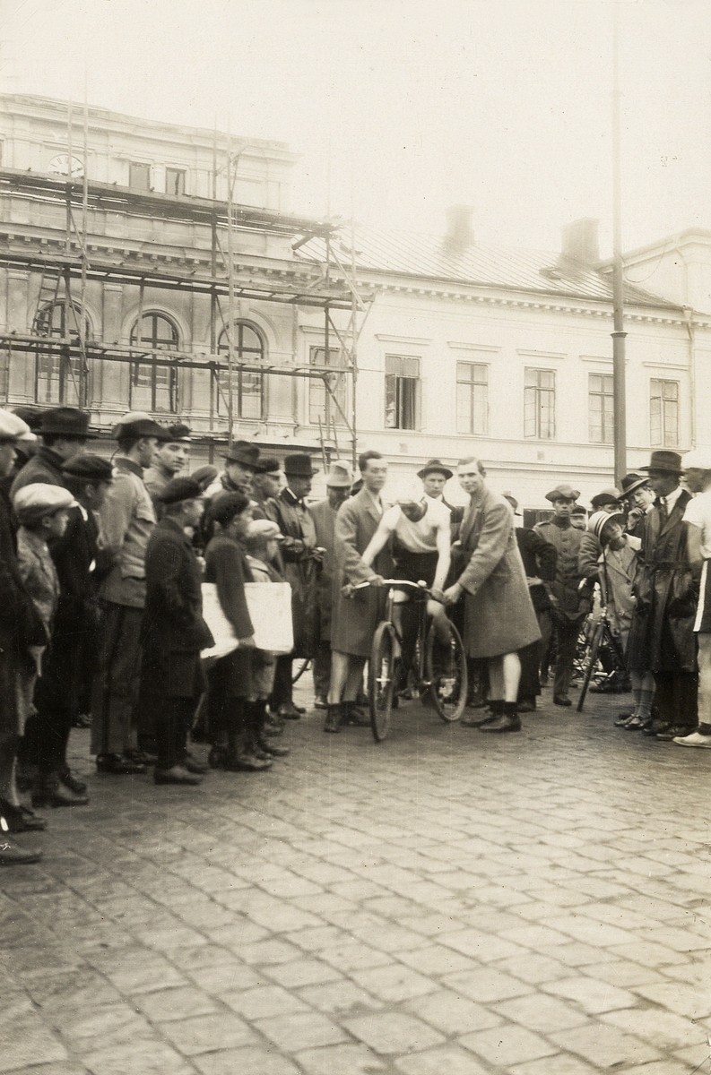 Cykeltävling på Stortorget, Växjö, ca 1925. En hel del nyfikna har samlats för att se starten.
I bakgrunden syns stadshotellet med byggnadsställning.