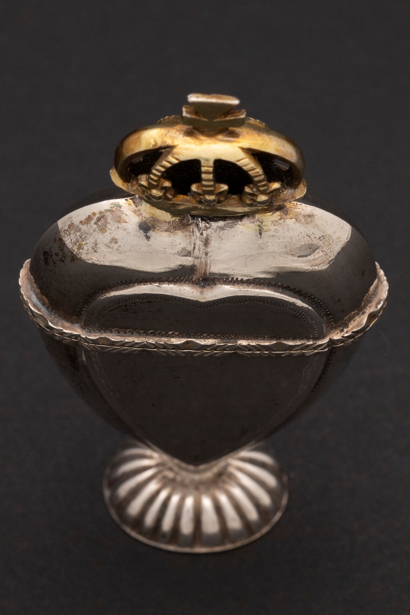Hjerteformet luktevannshus i sølv med oval fot og forgylt krone på lokket. Hjerteformede felt på for- og bakside av korpus. Enkel dekor. Innvendig forgylt.