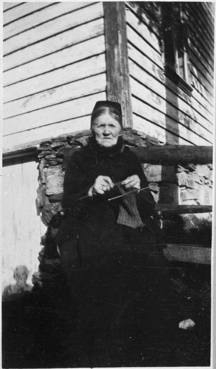 Inger Nilsdotter Lunde strikker i finvêret på gardstunet på Lunde i Ølensvåg, ca. 1925. Ho var omlag 90 år på dette biletet.