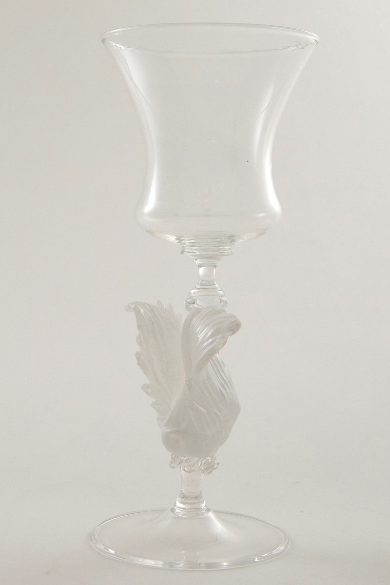 Glasspokal med kalkformet kupa. Stetten er utformet som en svane i opakt glass, og hviler på en sirkulær fot. Utformingen henspiller på venetianske vingeglass fra 1600-tallet, evt. nordeuropeiske glass utført i "façon de Venise".