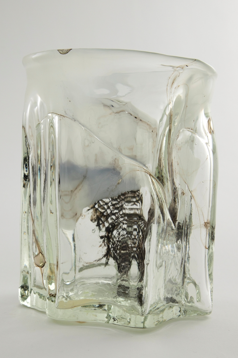 Tilnærmet sylindrisk vase i klart glass med innlagt dekor. Vasens tykke vegger har bølgende former og avsluttes i en sirkulær irregulær munningsrand. Innslag av melkehvite partier i opalinglass, samt fiberduk og luftblærer.
