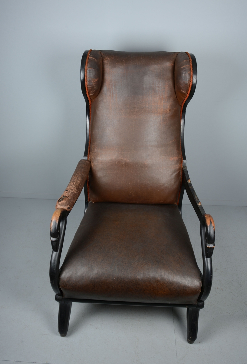 Ørelappstol med åpne, polstrede armlener, med støtter formet som svanehoder. Agramanbånd langs sidene på ryggen. Fjæring i sete.