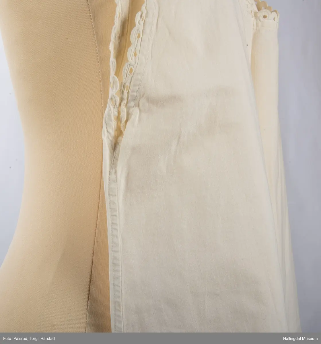 Hvit nattkjole i bomull med hvite blonder rundt hals og ermer. En hvit og en grå metallknapp på hver sin skulderstropp holder kjolen oppe.
