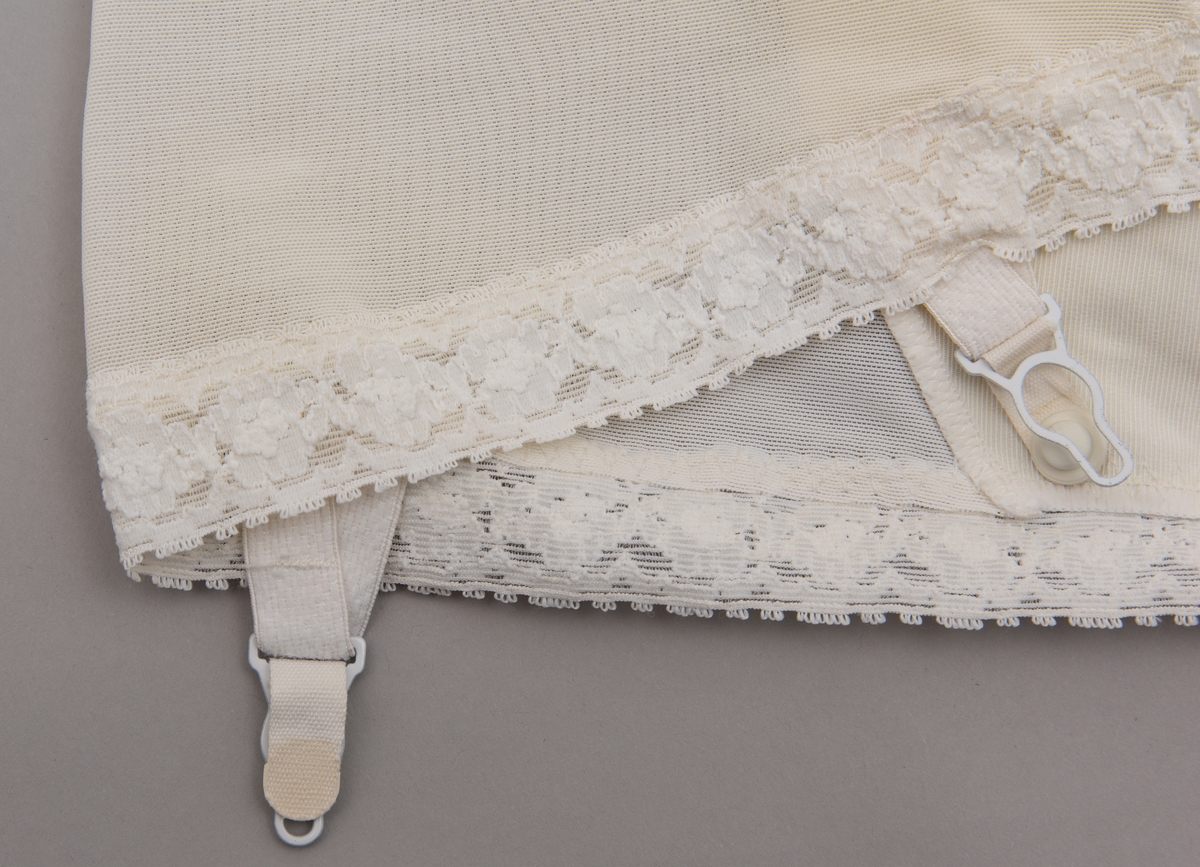 Hvit hofteholder med stropper og blondekant nederst og foran på midtstykke. Pyntet med sløyfe og kulørt blomst i tekstil foran på linning.