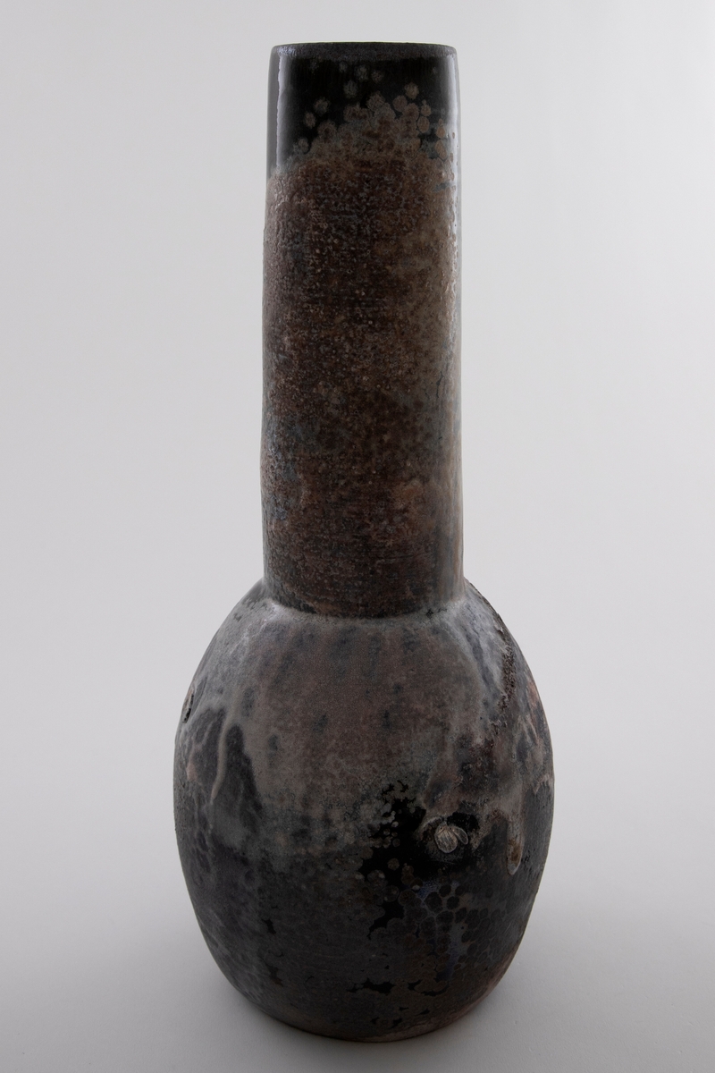Vase i glasert keramikk. Polykrom glasur i brune og svarte nyanser med rennende hvit begitning. Liten rund kolbe med lang hals.