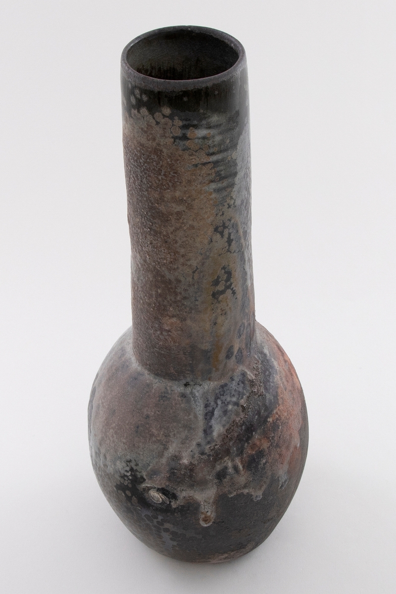 Vase i glasert keramikk. Polykrom glasur i brune og svarte nyanser med rennende hvit begitning. Liten rund kolbe med lang hals.