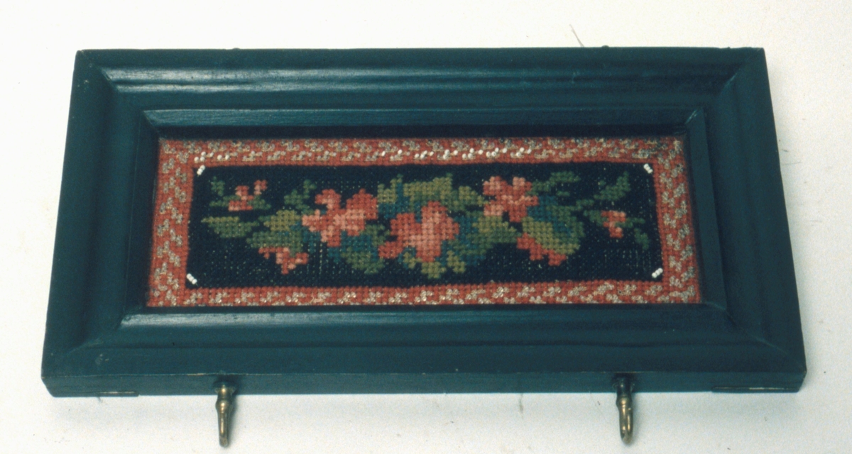 Handdukshängare, (hållare). Korssömsbroderat blommönster i rött, grönt, blått och svart samt vita glaspärlor på stramalj. Svartmålad träram med två ringar för upphängning och två krokar för handdukar.