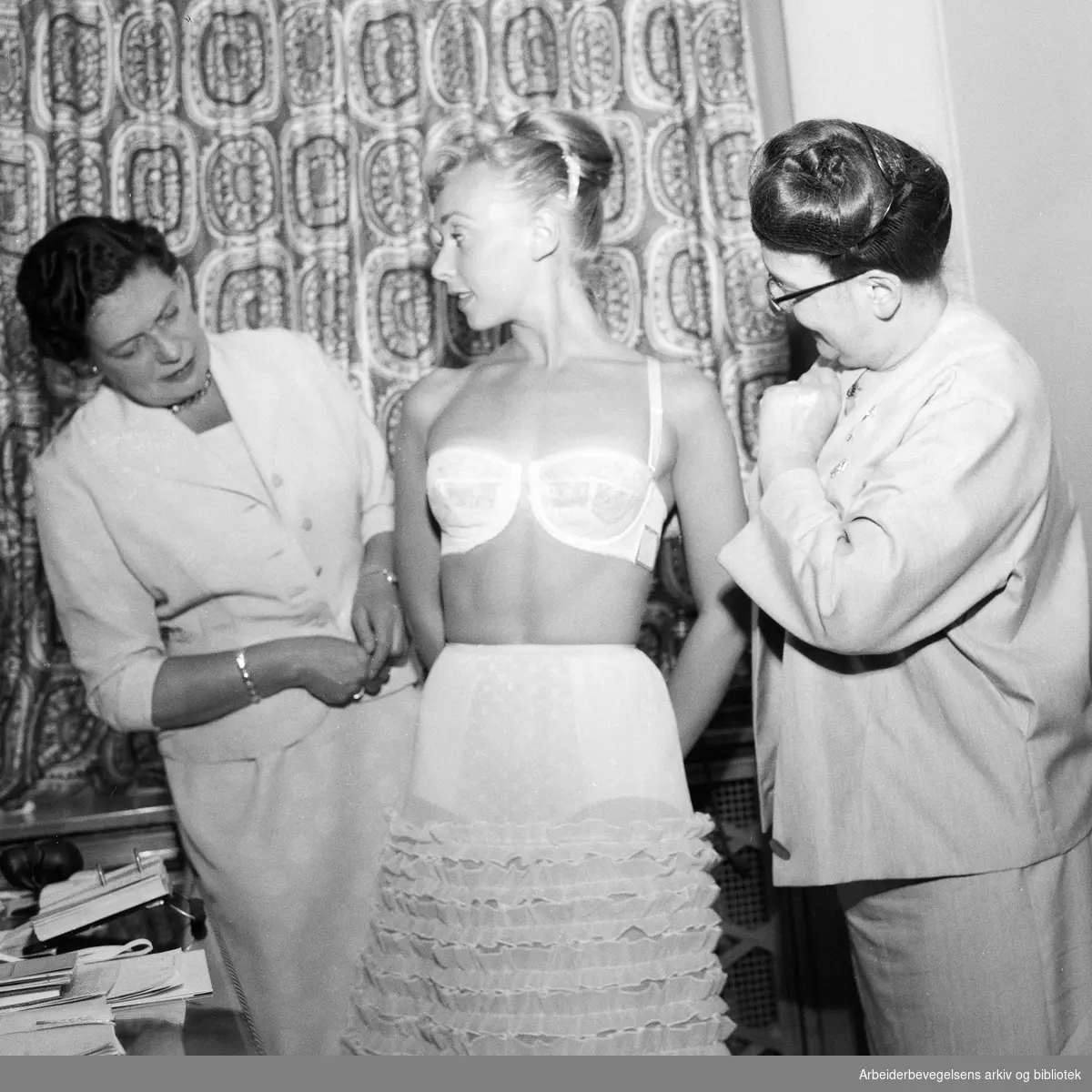 Korsett-eksperten miss Greaway, generalsekretær i Corset Guild of Great Britain, på besøk i Oslo. Til høyre, konsulent fru Irene Priestley-Røstad. Irene Nevermann sto modell. Juli 1957.