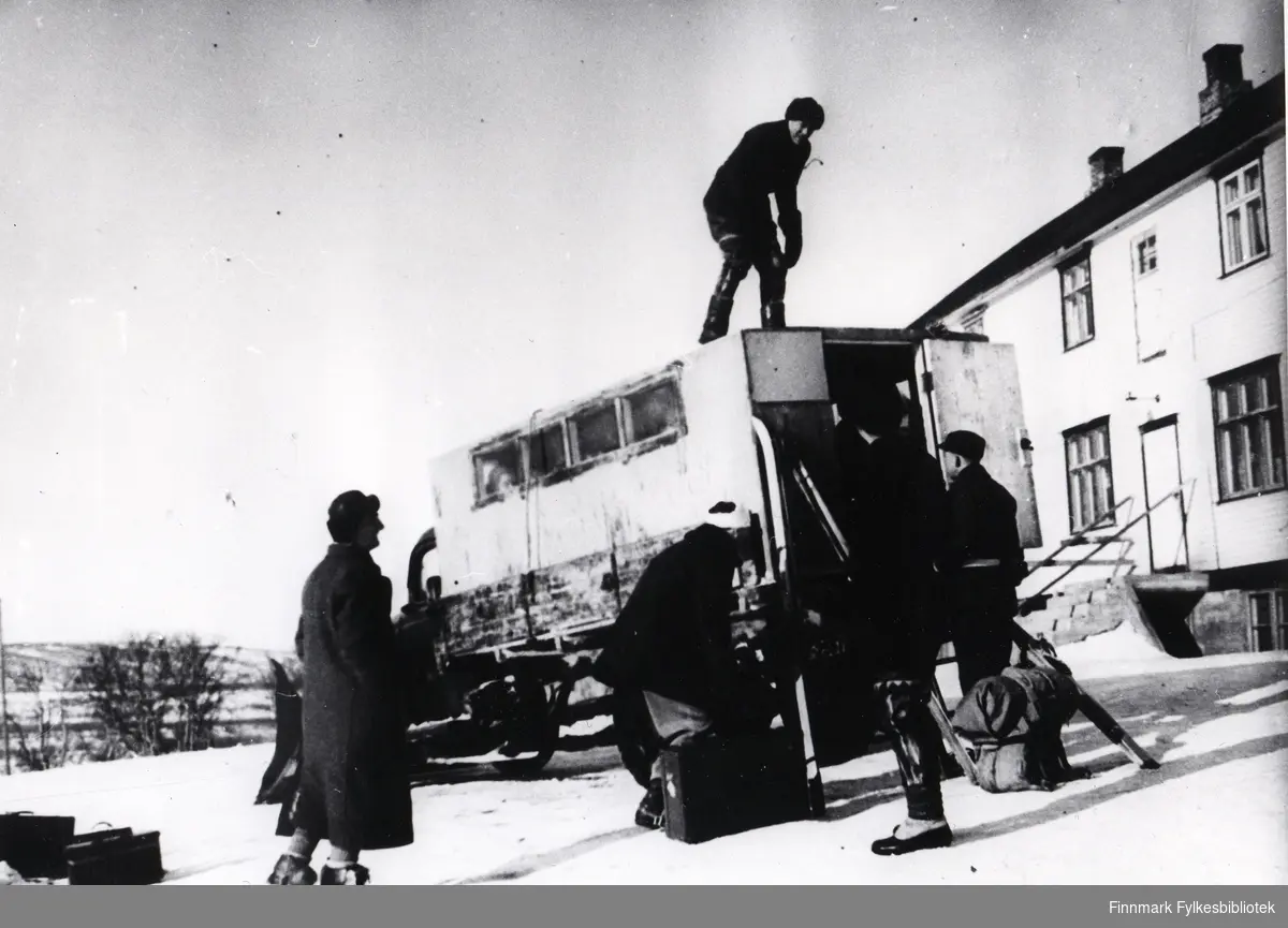 Vinteren 1946/1947 var det ikke noen veiforbindelse mellom Øst- og Vest-Finnmark. Veisjef Hofseth som kjente til forholdene på Tana elva fikk Jakob Nordgard til å kjøre brøytebilen på isen fra Skiippagurra til Karasjok. Bildet viser forberedelsene. Polarbil bussrute, Tanaelva. Passasjerer venter utenfor bilen. 