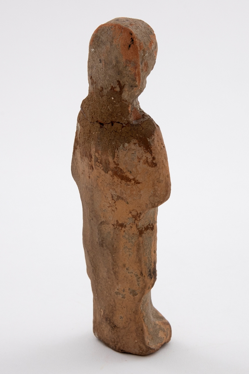Fragment av figurin i terrakotta. Lett stilisert formspråk. Fremstiller en kvinne i helfigur, iført drakt og med hodeplagg. Hul på innsiden. Enkelte synlige brudd og rester av jord.