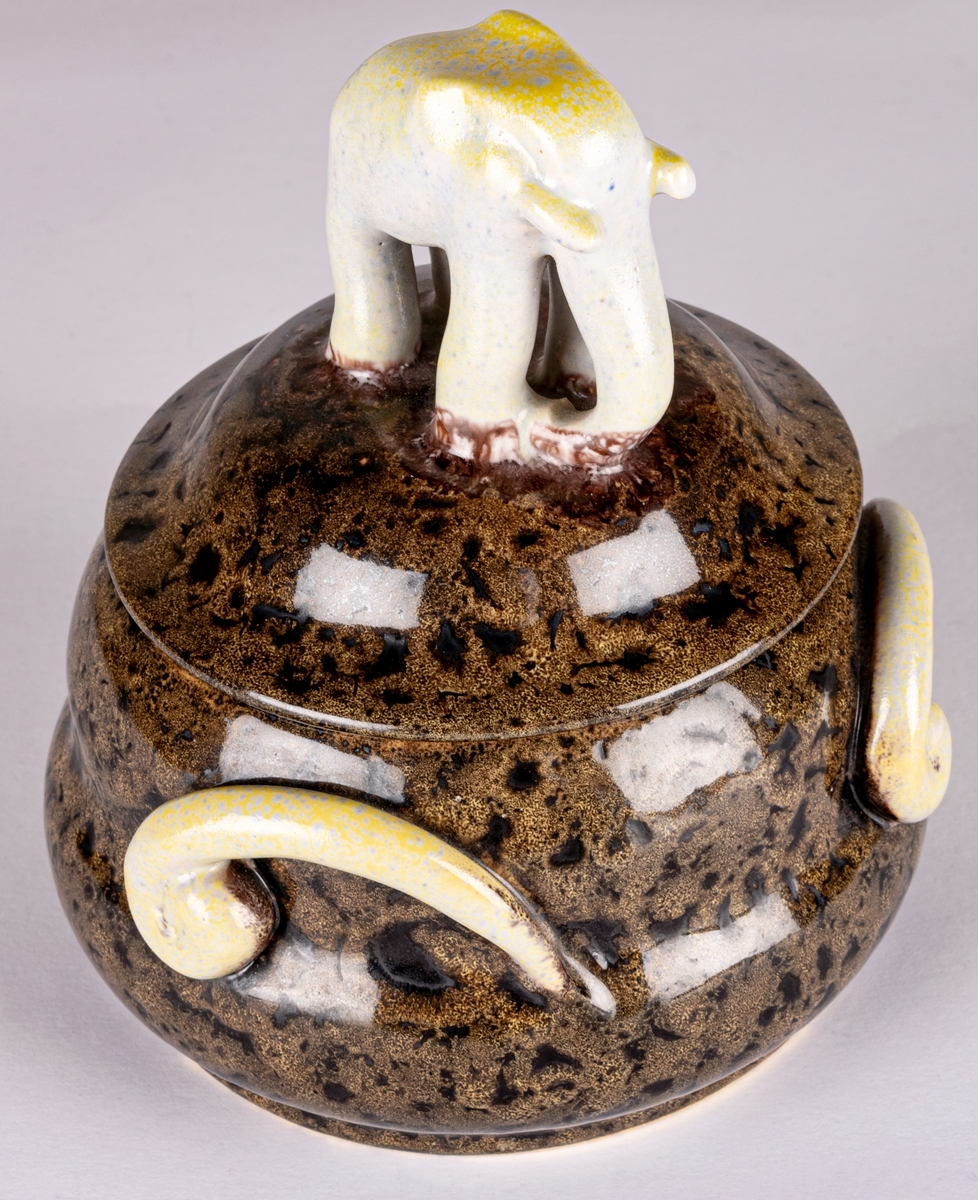 Bonbonjär, konfektskål med lock, formgiven av Allan Ebeling. Brunspräcklig glasyr, tre dekorationsöron på sidorna i en gulskimrande glasyr, locket kröns av en modellerad elefant i en gråblå och gulskimrande glasyr.