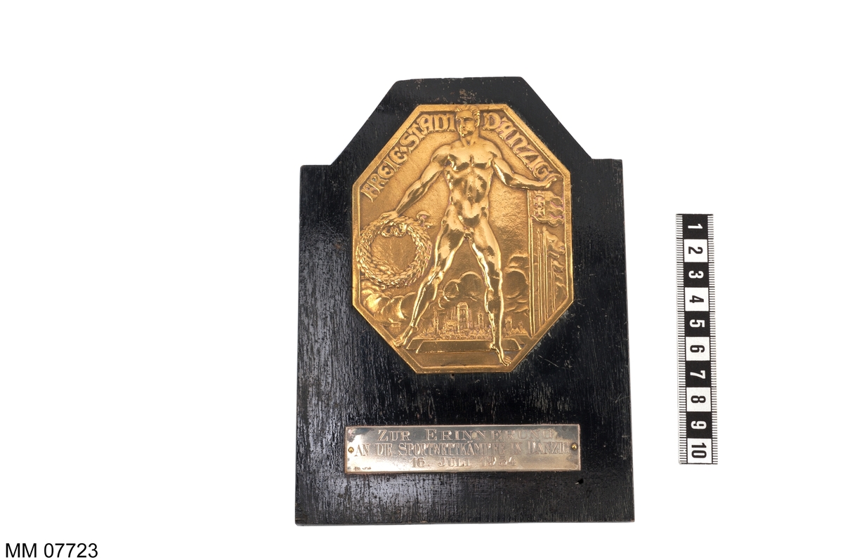 Plakett av brons på platta av trä, svart.
Utgör minnespris, erövrat i Dantzig 1934.
Inskription: Freie Stadt Dantzig.
På platta av silver: Zur Erinnerung An Die Sportwettkampfe In Dantzig 16. Juli 1934.
