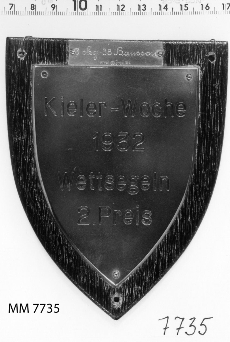 Plakett av brons på platta av trä, svart.
Inskription: Kieler-Woche 1932 Wettsegeln 2.Preis.
På silverplåt: 3 skg. 38 Hansson.