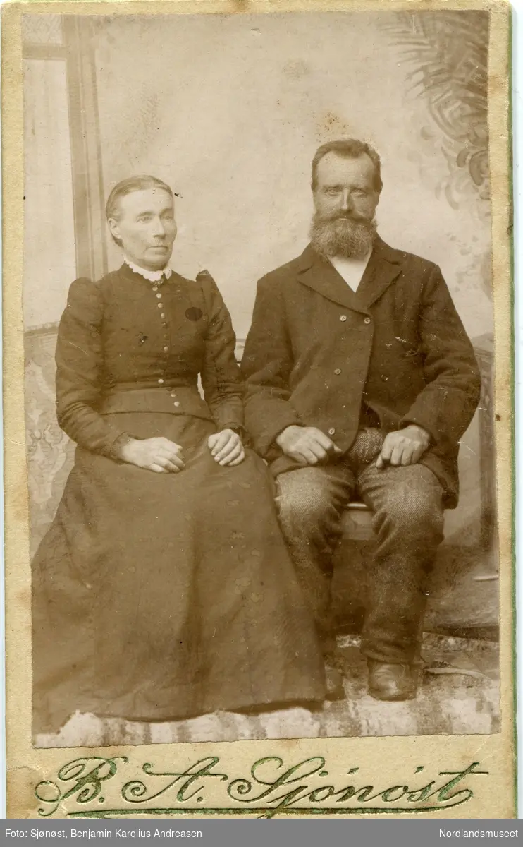 Portrett av et par, mann og kvinne. Norman Iversen f. 15.10.1842 og Anna Marie Bottolfsdtr f. 9.8.1840,  i et improvisert fotostudio, fotografens lerret litt skjevt.