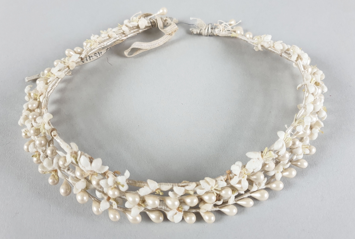 Kort, beige halsbånd bestående av fire kjeder besatt med kunstige perler og stoffblomster. Festes med strikk i nakken.