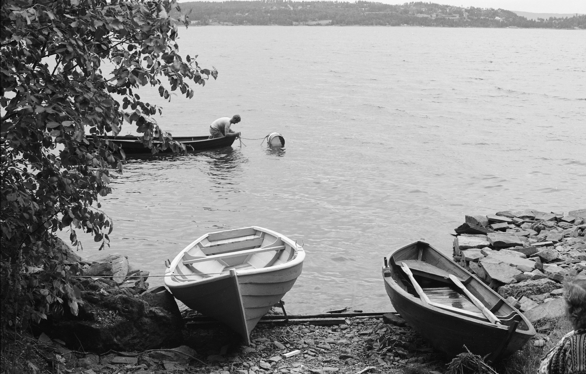 Fra båtstøa til yrkesfisker Paul Stensæter (1900-1982) ved Steinsfjorden, en sidearm til Tyrifjorden på Ringerike i Buskerud. På stranda i forgrunnen ser vi to robåter som fiskeren disponerte. Ute på vannspeilet lå en tredje båt, som var fortøyd i ei flytende tønne som må ha vært forankret på botnen av fjorden.