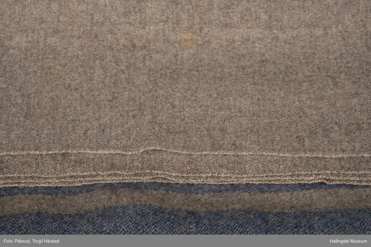 Grå kvitil, eller sengeteppe (ullpledd) med en bred og to smalere, blå striper midt på. 1-tråds renning og 1 tråds innlag.