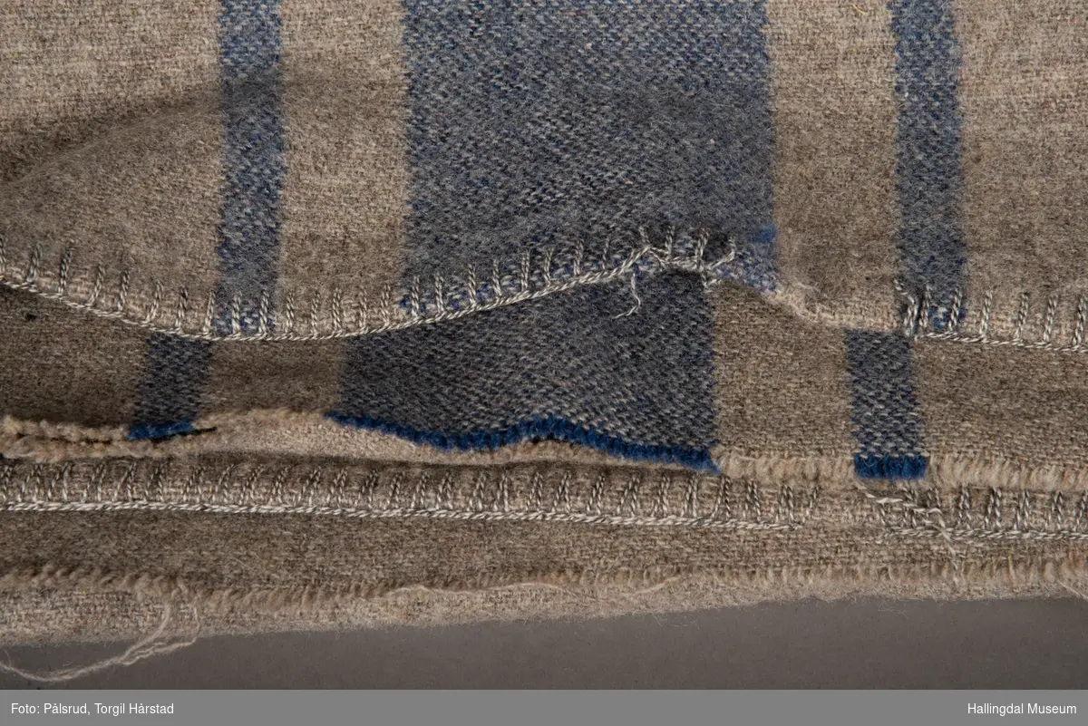 Grå kvitil, eller sengeteppe (ullpledd) med en bred og to smalere, blå striper midt på. 1-tråds renning og 1 tråds innlag.
