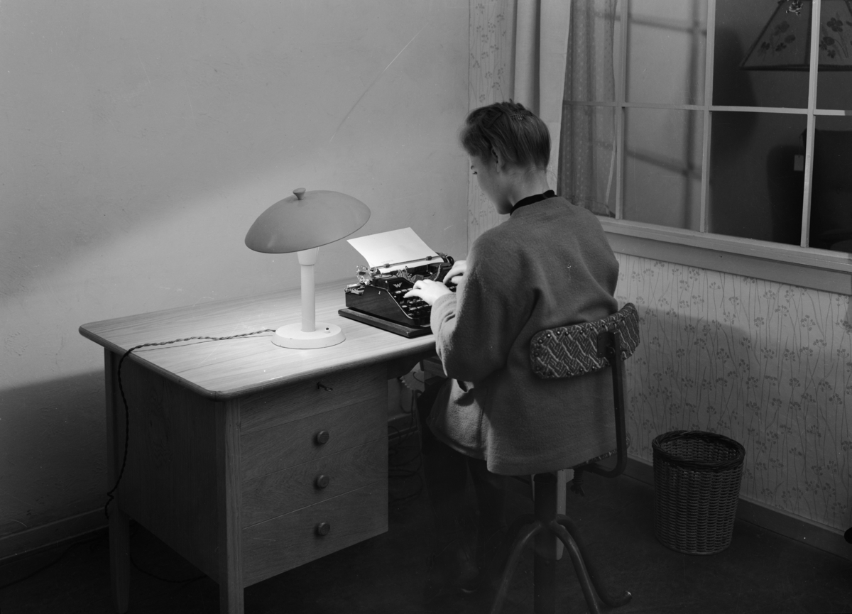 Produktfotografi, P,C.P. bordlampe. En kvinne sitter med ryggen til på en kontorstol ved et skrivebord og skriver på maskin. Ved siden står det en flettet papirkurv på gulvet. Til høyre er det et vindu med gardiner og en vegg med blomstrete tapet. Bordlampen lyser opp det halvmørke rommet. Publisert i Bonytt, januar 1949.