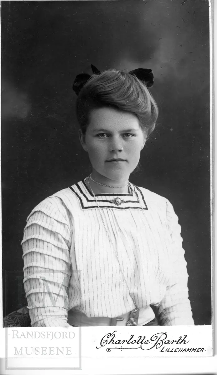 Brystbilde av ung kvinne i lys bluse med smale mørke striper. Mørk sløyfe i håret, langt halskjede og en liten brosje i halsen.