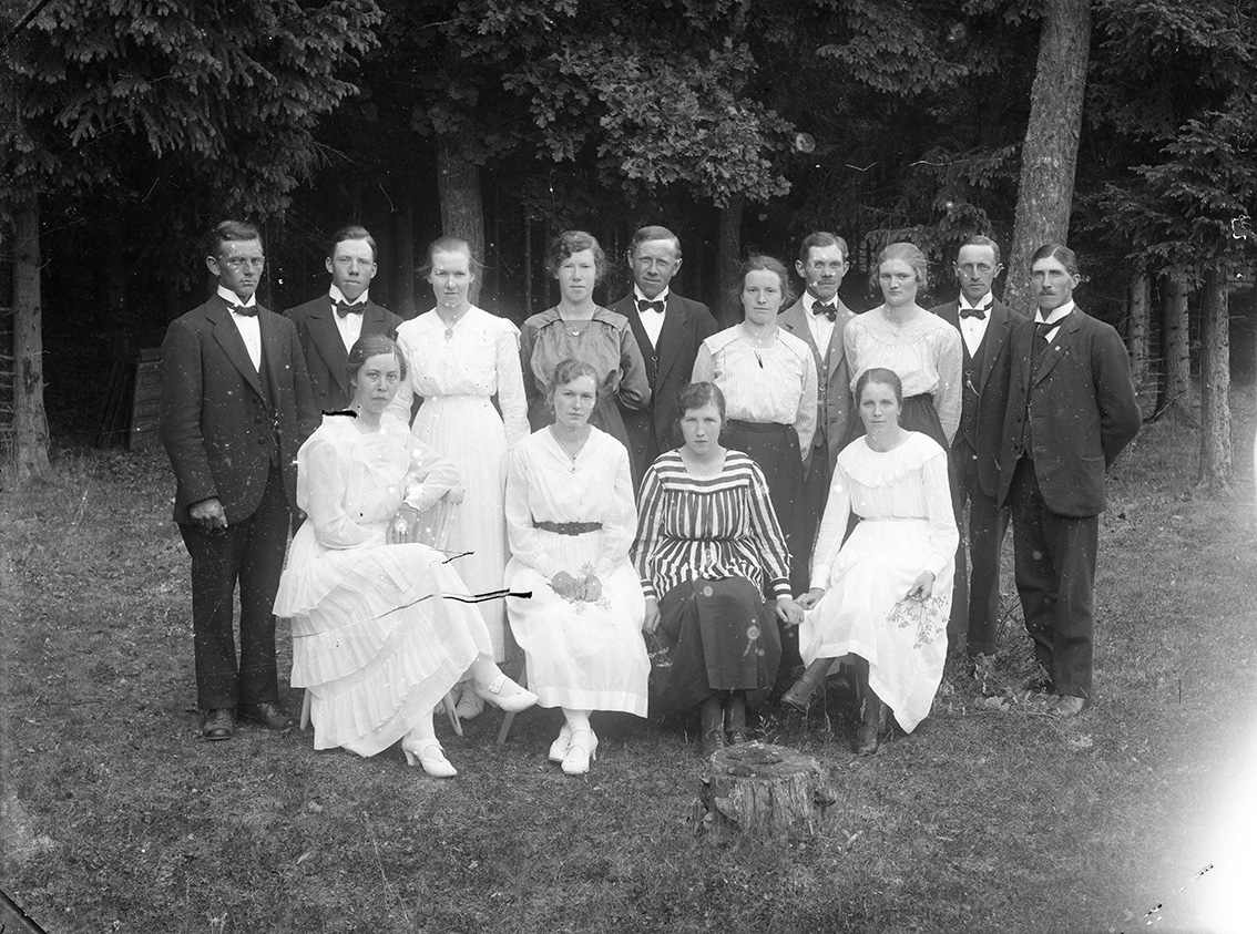 Gruppbild med 14 unga kvinnor och män sitter och står framför ett skogsparti.