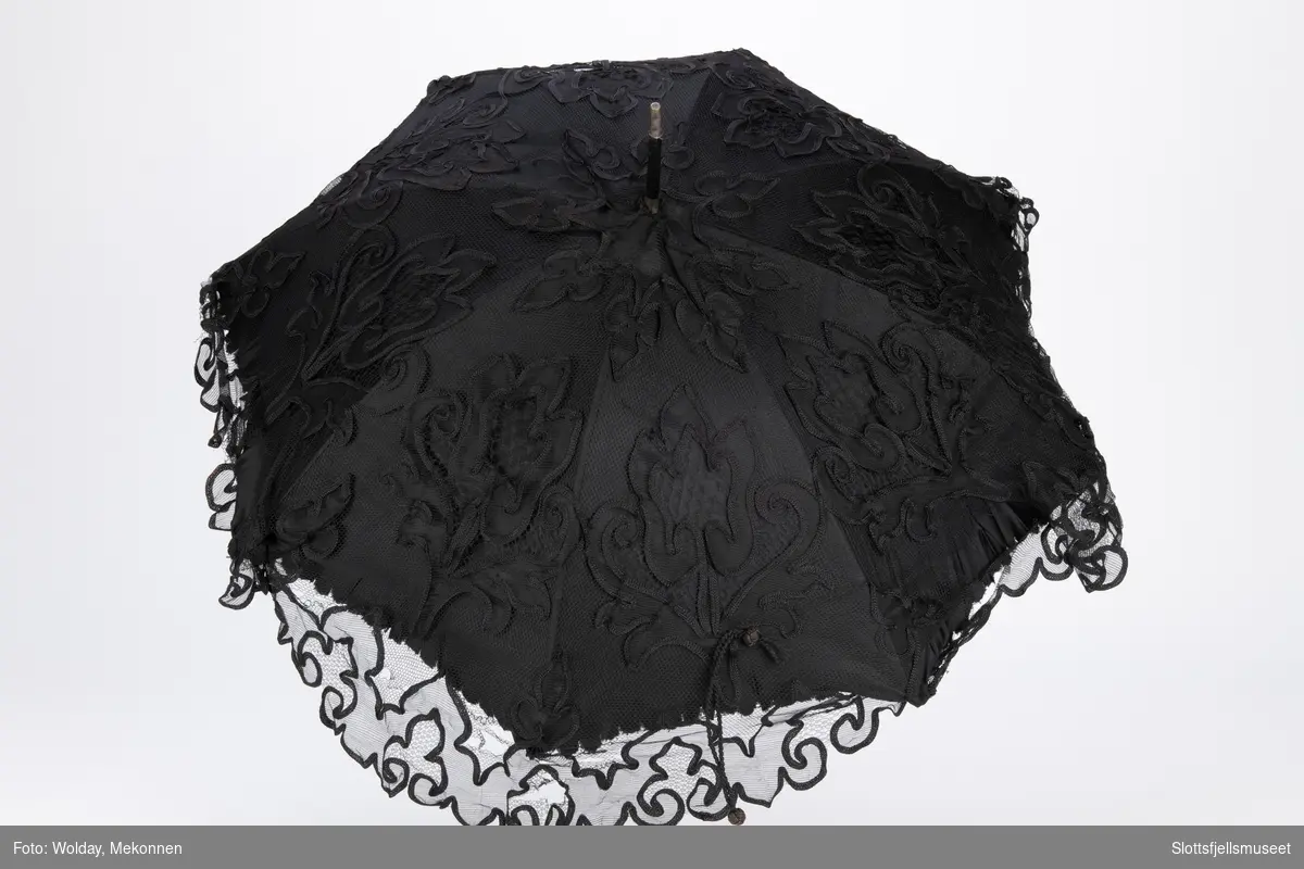 Parasoll med 8 spiler av metall og treskaft. Parasoll er trukket med svart stoff og en utenpå liggende svart tyll med dekor.