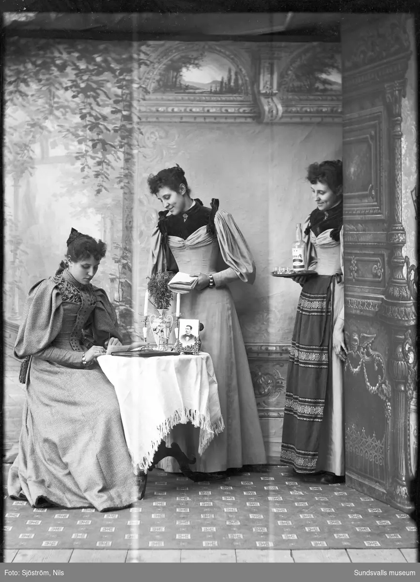 Ateljéporträtt med tre unga kvinnor, möjligen systrar. Arrangerad bild där en av dem sitter vid ett litet bord och skriver brev, på bordet står ett foto av en man.