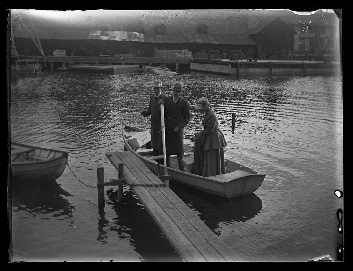 Två män och en kvinna i roddbåt invid brygga. Stockholm.