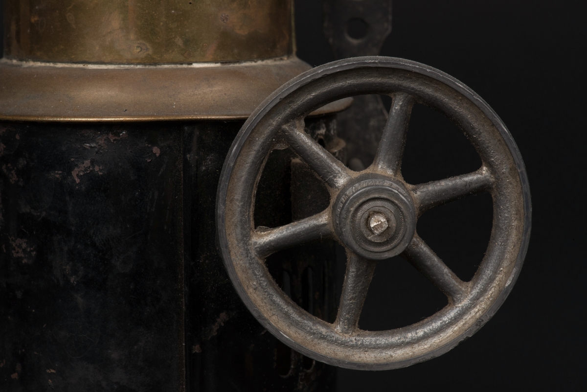 Ångmaskin av mässing och svartmålad plåt. 
Skorstensformad ångmaskin med hjul- och drivaxel på sidan. 
I bottendelen eldstad, sedan skorsten och överst en ventil.