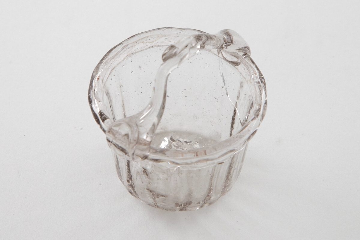 Kurvformet skål i klart glass med en grålig tone. Sirkulært korpus med vertikale riller. En svakt bøyd glasstav med løkkke fungerer som hank. Puntemerke på undersiden av skålen.